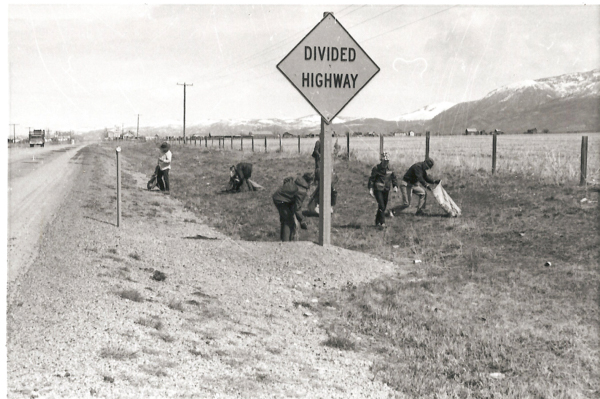 A group of people picking up trash beside a road. They are working near a sign that reads "DIVIDED HIGHWAY." The setting includes grassy fields and mountains in the background. A fence runs alongside the road, and a vehicle is visible in the distance.