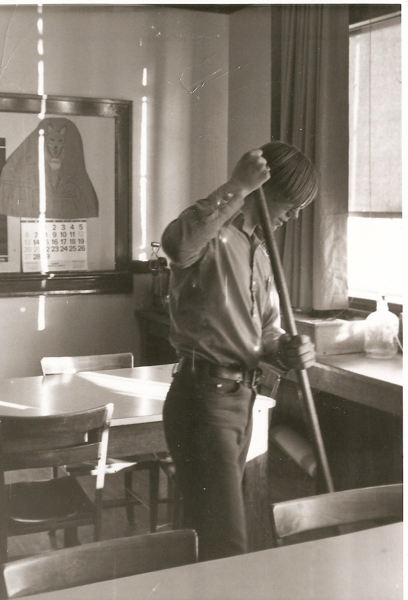 A person sweeping the floor in a room with tables and chairs. A calendar with numbers and an image of a fox or similar animal is visible on the wall. There is a large window with curtains in the background.