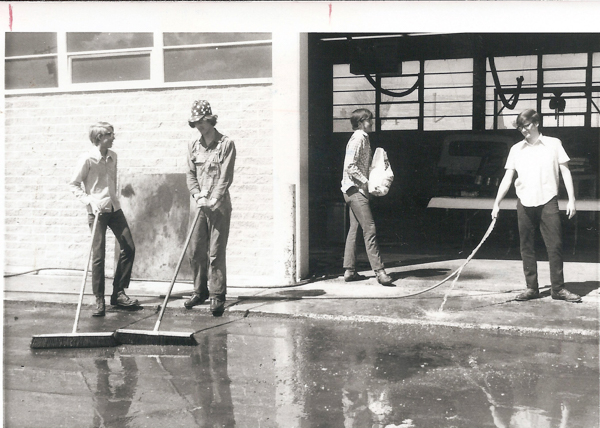 Four people are standing outside a building with a large door. Two are using push brooms to sweep water on the ground. Another person is carrying a bag, and the fourth is holding a hose, spraying water. They are in front of a windowed wall, and inside the building, part of a vehicle is visible.