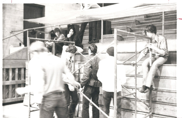 A group of people are gathered around scaffolding. Two individuals are sitting on the structure, one on the right holding a piece of it, while several others stand or sit around it. The setting appears to be an outdoor area with steps or seats in the background. Some people are interacting while others are observing.