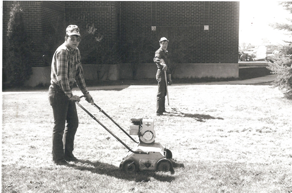 Two men are working on a lawn. One man is pushing a lawn aerator in the foreground, wearing a plaid shirt and cap. The other man, standing in the background, holds a garden tool and is wearing a work outfit and cap. They are near a brick building.