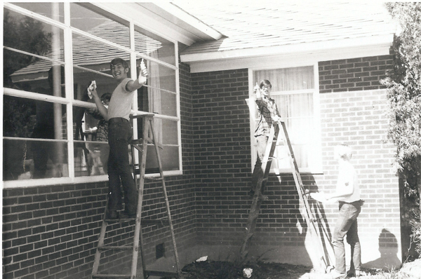 Three people cleaning windows on the exterior of a brick building. Two individuals are standing on ladders, each holding cleaning supplies and smiling towards the viewer. The third person stands nearby on the ground, also holding cleaning materials.