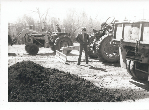 A person stands next to a tractor and a dump truck at a construction site. Another person is seated on a tractor labeled "International 185." The ground is covered in dirt piles, and there are leafless trees in the background. A sign on a plow blade reads "BIG RHINO."