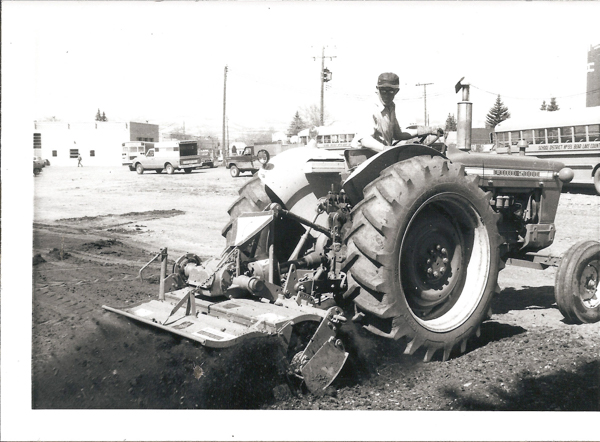 A person driving a large tractor on a dirt surface. The tractor has a plow attachment, and there are buildings, vehicles, and school buses in the background. The text on the bus reads "SCHOOL DISTRICT #33 BEAR LAKE COUNTY." The tractor has the words "FORD 5000" visible on the side.