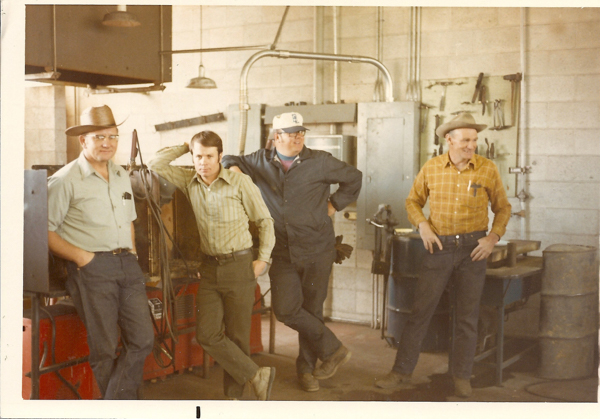 Four men are standing in a workshop. The first man on the left wears a hat and glasses, with his hands in his pockets. The second man leans against a piece of equipment and has his hand on his head. The third man wears a cap, glasses, and work clothes, with one hand on his hip. The fourth man on the right, also wearing a hat, looks to the side and has his hands in his pockets. The background includes tools on the wall, metal barrels, and industrial equipment.