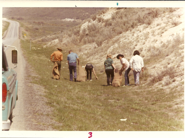 A group of people standing near the side of a road, with a hillside on the opposite side. They appear to be picking up something from the ground. One person is bending over, while others are holding or carrying large bags. A truck is parked on the roadside. The number "3" is written at the bottom of the image.