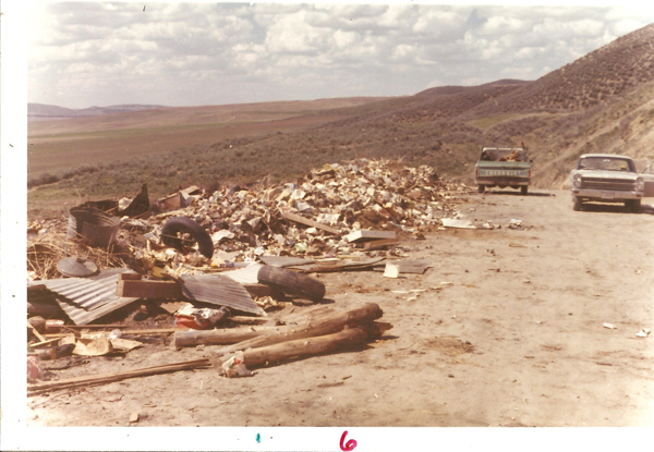 A dirt road with two parked trucks, one open-bed and the other enclosed. Piles of debris and garbage, including tires and metal sheets, are scattered along the roadside. The background features rolling hills and an open sky with clouds. At the bottom of the image, the number "6" is written.