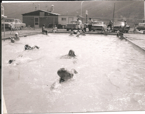 People swimming in a rectangular outdoor swimming pool. Several swimmers are in the water, while others sit or stand around the pool's edge. In the background, there is a chain-link fence, parked cars, and a building. Mountains are visible in the distance.