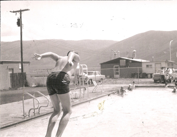 A person, wearing swim trunks, is seen in mid-motion preparing to dive into a swimming pool. In the background, several people are sitting or standing near the pool. There is a building, vehicles, and a fence, with mountains visible in the distance.