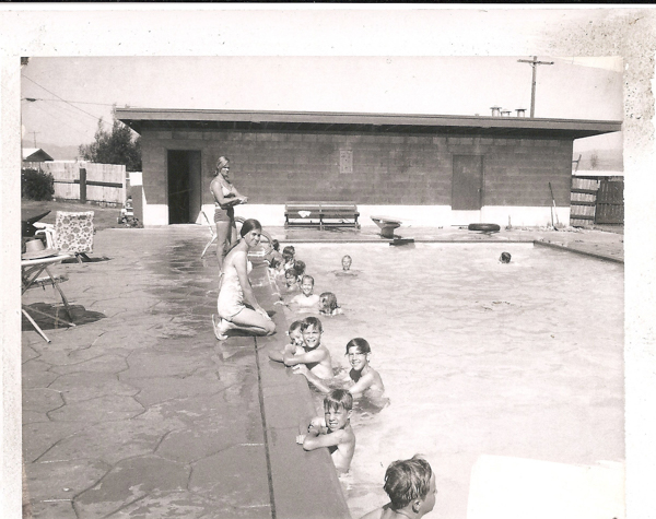 A group of children is swimming in a pool, with a few sitting on the edge. A woman kneels at the poolside, watching the children. Another person stands near a building in the background. There are lounge chairs and a bench around the pool area. The setting appears to be a recreational or community swimming pool.