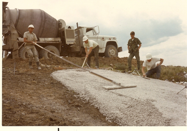 Four men working on a construction site with a truck in the background. Two men are spreading concrete using tools, one is smoothing the surface, and the other stands nearby observing. Some are wearing hard hats and one is in a military-style uniform. The truck has the word "CHALLENGE" visible on its side.