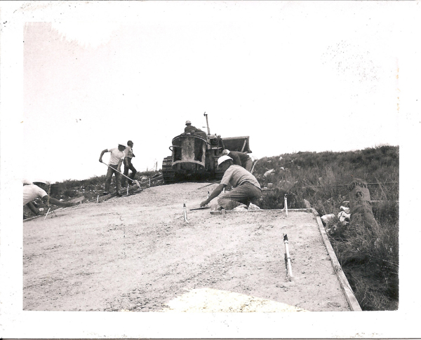 Men are smoothing a newly laid concrete surface on a sloped path, using tools. A large machine is positioned in the background, with two men standing nearby. A bag labeled "ASPHALT" is visible on the right side amidst grass.