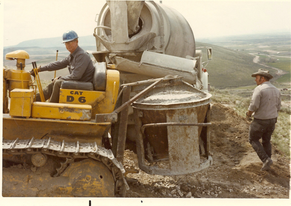 A man wearing a blue hard hat is seated on a yellow Caterpillar D6 bulldozer. Another man in a cowboy hat stands nearby, observing. They are working with a large cement mixer on a hilly construction site with a view of a valley and distant landscape in the background.