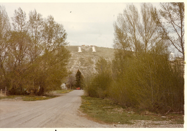 A dirt road leading towards a hill with large letter "M" displayed on its slope. Tall trees flank the road, and there is a small building and a parked vehicle near the base of the hill.