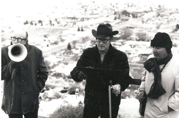 Three men are standing on a hillside. The man on the left holds a megaphone and is wearing glasses and a coat. The man in the center is wearing a hat and glasses and is holding a piece of paper on a clipboard. The man on the right is wearing a coat, a hat, and a scarf. The background shows an expansive view of a town.