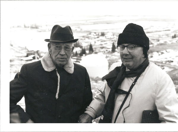 Two men standing outdoors with a snowy landscape in the background. The man on the left wears a coat with a fur collar and a hat, while the man on the right is dressed in a jacket, hat, and scarf, and holds a camera around his neck.