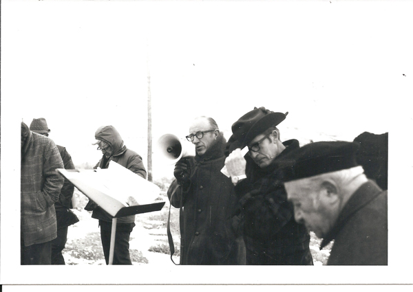 A group of men are standing outdoors. One man is holding a megaphone, wearing glasses and a coat. Another man is adjusting his hat, and a third man is looking down with a cap on his head. A person in a checkered coat is looking at a document on a stand. The scene appears to be in cold weather.