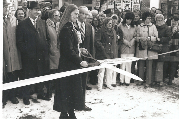 A group of people is gathered outdoors for a ribbon-cutting ceremony. A woman with long hair, adorned with a flower necklace, holds a pair of large scissors to cut the ribbon. Several people stand in the background, wearing coats and hats. There are signs in the background displaying prices and promotions for items like grapefruit and flour.