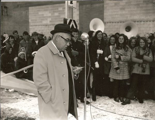 A man wearing a coat and hat stands at a microphone, reading from a piece of paper. Behind him, a group of people, including some holding musical instruments like clarinets and tubas, stand together. The ground is covered in snow, and a building is visible in the background.