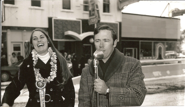 A man and a woman stand outdoors on a street. The man is holding a microphone and speaking, wearing a plaid coat. The woman is smiling broadly, wearing a coat with a floral lei around her neck and a sash that reads "Miss Idaho," along with the number "71." Buildings and people are visible in the background.