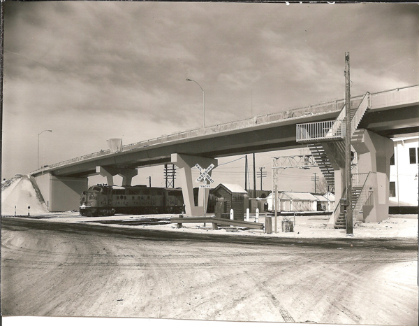 A train labeled "Union Pacific" travels beneath a large overpass. The area is snowy with visible tracks. To the right, there's a staircase leading up to the bridge. A railroad crossing sign reads "Railroad Crossing 5 Tracks." In the background, there are several utility poles and buildings.