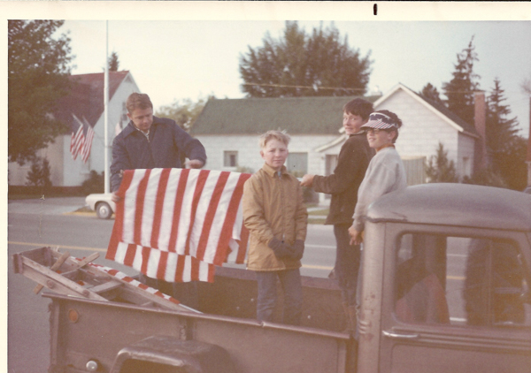 A group of four people stand in the back of a pickup truck. One person is holding a large flag with red and white stripes. They appear to be outdoors, with houses and trees in the background. An American flag is visible in the distance, next to a building.