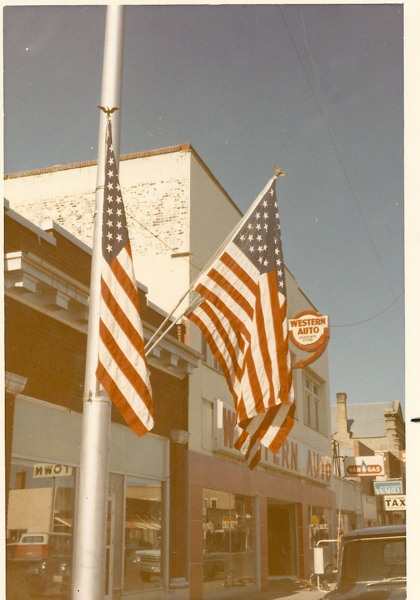Two American flags are displayed on poles in front of a storefront. The building features signage that reads "WESTERN AUTO" and a circular sign with "WESTERN AUTO ASSOCIATE STORE." Additional signs in the background indicate "VAN & GAS" and "TAX." Part of a vehicle is visible near the bottom right corner.