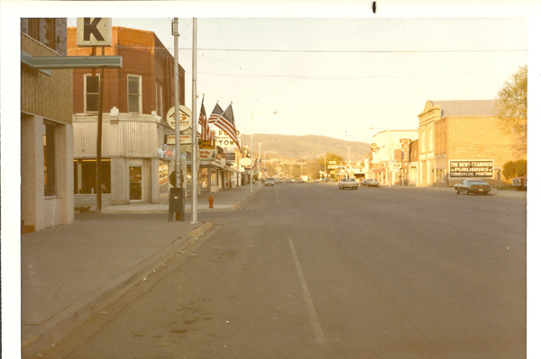 A deserted street scene with buildings on both sides. On the left, there is a sign with the letter "K" and a building with a sign for a "TAX" service. Further down, there are American flags and a sign that reads "FOOD TOWN." On the right side, a sign reads "THE NEWS-EXAMINER PUBLISHERS & COMMERCIAL PRINTING." A few cars are parked along the street, and hills are visible in the background.