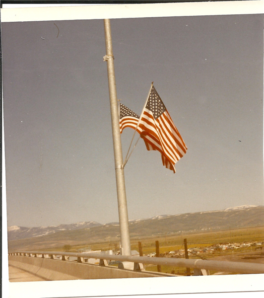 Two American flags on a pole, slightly tilted, with a landscape of mountains and fields in the background. A fence and a bridge railing are visible in the foreground.