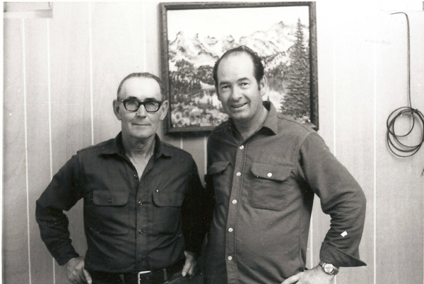 Two men standing side by side in a room. One man is wearing glasses and both are wearing long-sleeved shirts with pockets. Behind them, there is a framed landscape painting of mountains and trees. A coiled wire is hanging on the wall to the right.