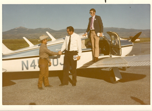 Two men are standing next to a small aircraft, shaking hands. One man is wearing a hat and casual clothing, while the other is in a shirt and tie. A third man, wearing a suit and holding a book, stands on the wing of the plane. The plane has "N4509J" written on its side. A mountainous landscape is visible in the background.