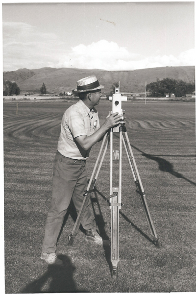 A man is standing on a grassy field using a theodolite mounted on a tripod. He is wearing a short-sleeved shirt, pants, and a straw hat. The background includes hills and trees with a few structures visible in the distance.