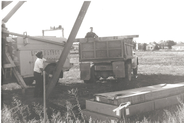 Two men are working in a field. One man stands beside a truck with a large chute, and another is in the back of a separate truck. The trucks are positioned on a grassy area with some buildings visible in the background. There is a stack of boxes on the ground nearby. The truck with the chute has partial text visible on it.