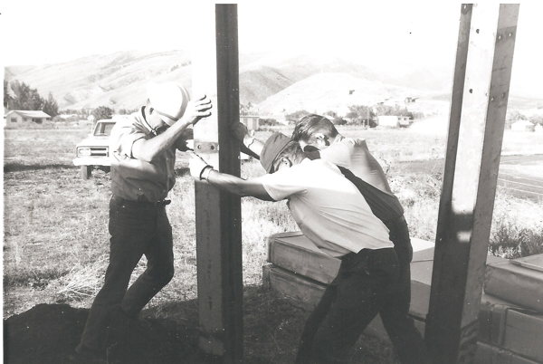 Three men are working together to install a large vertical beam in a grassy open area. They are pushing against it, appearing to adjust or secure its position. One man is wearing a hard hat. In the background, there's a truck and some buildings with hills in the distance.