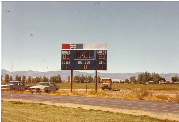 A scoreboard in an open field showing "Home 00," "Guest 00," "Down 1," "Yds. to go 10," and "Qtr. 1." A Pepsi logo is at the top of the board. Below, a track is visible with two parked cars and a yellow school bus. Trees, mountains, and a few buildings are in the background.