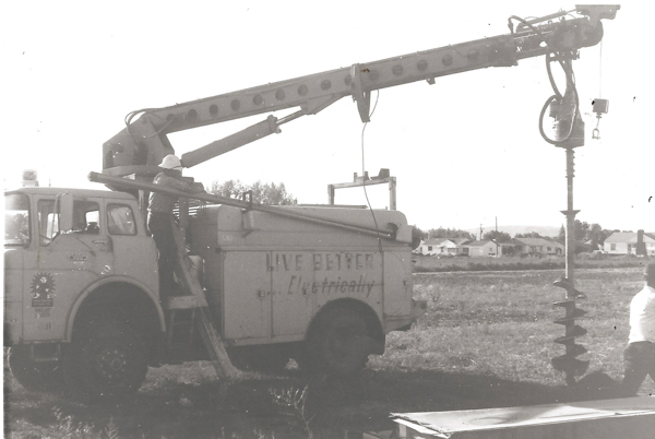 A truck equipped with a crane and auger stands in a field. A person wearing a hard hat is climbing onto the truck. The side of the truck has the text "LIVE BETTER Electrically." Houses and trees are visible in the background.