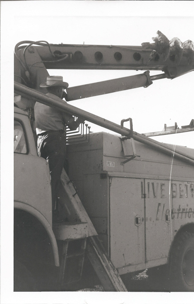 A person wearing a hat is standing on a step and working on the side of a utility truck. The truck has an attached crane with an auger. A portion of text on the truck reads "LIVE BET... Electri..." and there is a label that says "PLUG 1210".
