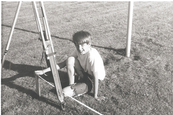 A young person sitting on grass next to a tripod. They are wearing a striped shirt and shorts. There is a string on the ground in front of them.