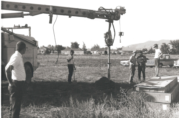 A group of five men stand in an open field next to a large piece of machinery. One man stands on a platform, while another holds a clipboard and talks to two men nearby. A fifth man stands a short distance away, examining the scene. In the background, there are houses, cars, and distant mountains. The side of a vehicle has the partial text "Electrical" visible.