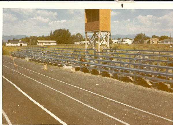 A set of metal bleachers next to a running track with a wooden tower nearby. There are a few people sitting on the bleachers. In the background, there are several small houses and trees, with mountains visible in the distance. The sky is partly cloudy.