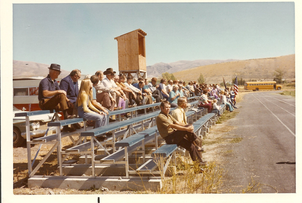 People seated on bleachers along a track, with a wooden structure above them. A school bus is visible in the background on a grassy area, and an American flag stands nearby. The scene is set in a rural, hilly landscape under a clear sky.
