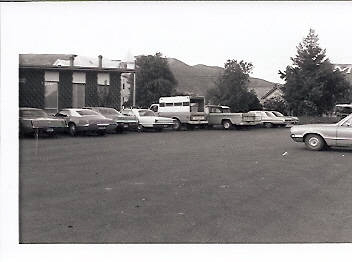 A row of parked cars lined up in a parking lot next to a building. In the background, there are trees and a mountain range. One car in the foreground is parked separately.