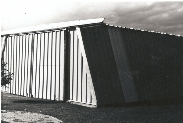 A large, angled metal structure with vertical panels stands on a grassy area. The structure appears to be divided into sections, with one section slightly darker than the other. The sky above is cloudy.