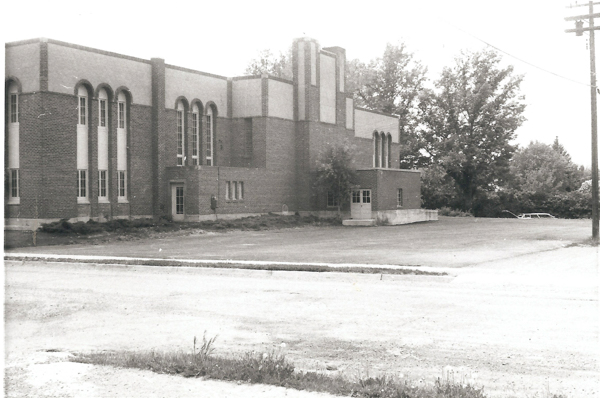 A large brick building with tall windows and arched designs. There are trees in the background and a parked car to the right. The foreground has a dirt road and some grass.