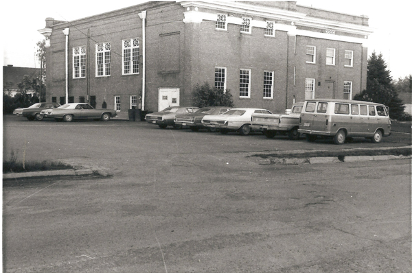 A parking lot with several cars and a van parked in front of a large brick building. The building has tall windows and a decorative roofline. The side of a Ford pickup truck can be seen, and there are some trees and shrubbery in the background.