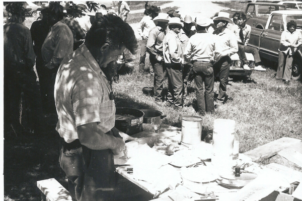 A man standing at a table covered with pots, pans, and various items, appears to be preparing or organizing something. In the background, a group of people are gathered, some wearing hats and engaging in conversation. Several cars are parked nearby, with a few individuals leaning against them. The scene is set outdoors on a grassy area.
