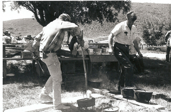 A man wearing a cap and plaid shirt is tending to a pot over a fire using a long stick, while another man in a short-sleeved shirt stands nearby, holding utensils. There are several pots and a wooden table nearby with supplies on it, including visible boxes with text that reads "Budweiser" and "Stoker's Sliced Beets." In the background, people are seated at a picnic table under a tree, and a car is parked further behind them.