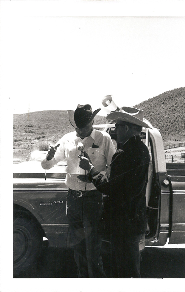 Two men wearing cowboy hats are standing next to a truck with an open door. One man is holding a microphone, and the other is gesturing with his hand. The truck has a visible emblem with the text "Custom/20." They are in a rural area with hills in the background.