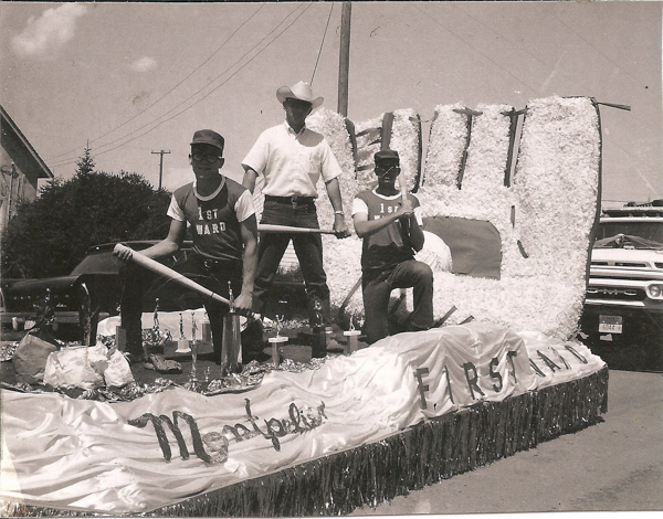 Three men on a parade float with a large baseball glove structure. Two men sit in front wearing "1st Ward" shirts, one holding a baseball bat, while a third man stands in the back wearing a cowboy hat. The float is decorated with fringes and has the words "Montpelier First Ward" on a banner. A GMC vehicle is visible in the background.