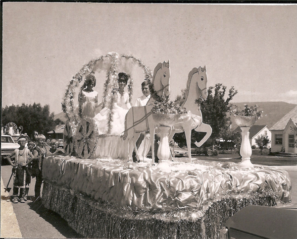 A festive parade float decorated with two large horse figures pulling a carriage, with three young women dressed in formal dresses seated inside. The float is adorned with shiny fabric and tinsel. A group of children dressed in costumes stand beside the float. In the background, there are houses, trees, and a partially visible car.